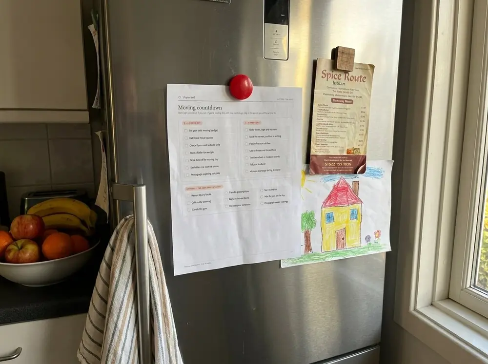 An Unpacked moving-countdown page held to the refrigerator with a magnet, next to a fruit bowl.